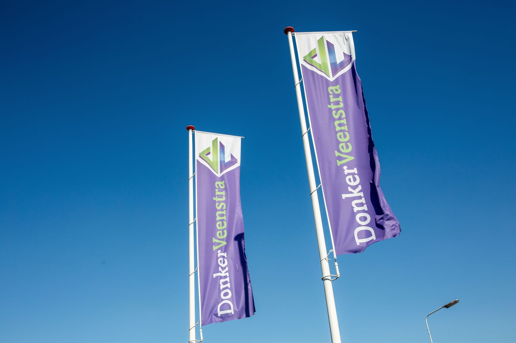 Twee paarse vlaggen met het bedrijf DonkerVeenstra erop. Bovenaan het groen en blauwe logo van DonkerVeenstra. Blauwe lucht als achtergrond.
